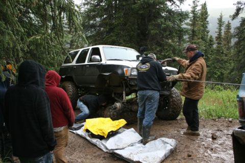 1990 Chevrolet Suburban - A group of people are working on the undercarriage of a lifted, two-tone (black and white) SUV that is stuck in the mud on a wooded trail.…
