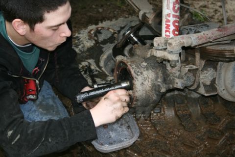 1990 Chevrolet Suburban - A person works on the front differential of a muddy vehicle, holding a greasy axle shaft. The scene appears to be outdoors with the vehicle…
