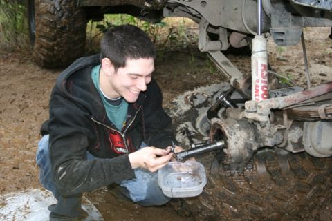 1990 Chevrolet Suburban - A young man kneels beside the front axle of a muddy off-road vehicle, working on a component with a small container of dark fluid. The vehi…