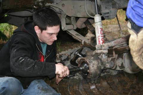 1990 Chevrolet Suburban - A person works on the front axle of a muddy off-road vehicle, likely a Chevrolet Suburban, with leaf springs and a shock absorber visible.