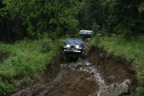 1990 Chevrolet Suburban - A blue Jeep Wrangler navigates a muddy, rocky trail through a dense forest. Another SUV is visible further up the trail.