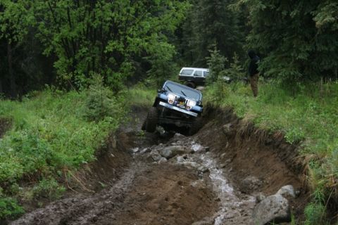 1990 Chevrolet Suburban - A blue Jeep Wrangler navigates a steep, muddy, and rocky off-road trail with lush green trees on either side. Another SUV is visible furthe…