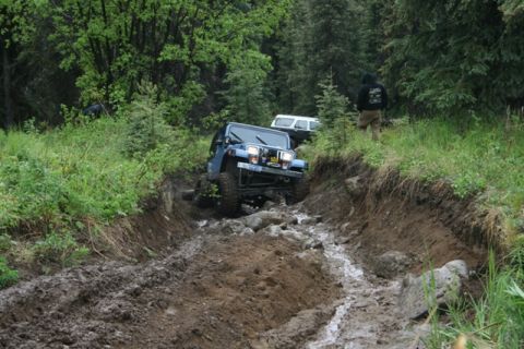 1990 Chevrolet Suburban - A blue Jeep Wrangler is navigating a very muddy and rocky off-road trail, with a white SUV visible behind it in the trees. A person in a bl…