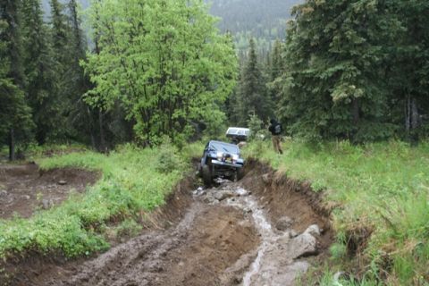 1990 Chevrolet Suburban - A blue Jeep Wrangler navigates a deep, muddy, and rocky off-road trail surrounded by lush green trees. Another vehicle, possibly a white SU…