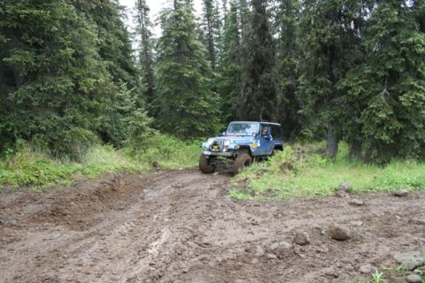 1990 Chevrolet Suburban - A blue Jeep Wrangler is driving on a muddy trail through a dense forest. The vehicle is angled towards the right, with its front tires deep…