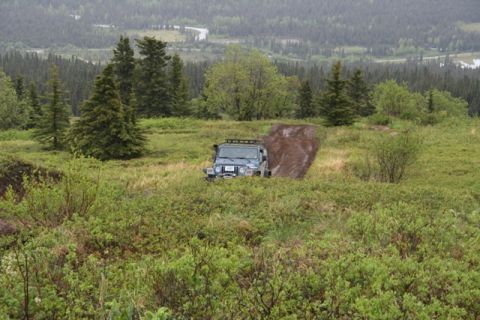 1990 Chevrolet Suburban - A blue Jeep Wrangler is navigating a muddy track on a grassy hillside. The vehicle is heading uphill, with dense green foliage in the foreg…
