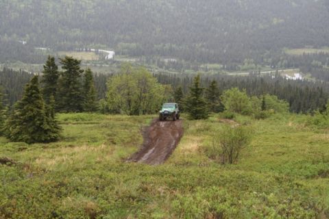 1990 Chevrolet Suburban - A green Jeep Wrangler drives up a muddy track on a grassy hillside, with a dense forest and a winding river visible in the background.