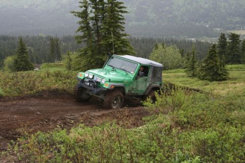 1990 Chevrolet Suburban - A green Jeep Wrangler is navigating a muddy, off-road trail with dense green trees and hills in the background.