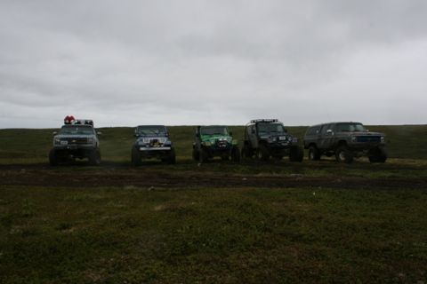 1990 Chevrolet Suburban - Five modified off-road vehicles are parked in a line on a grassy, uneven terrain under a cloudy sky. The vehicles include a light blue pick…