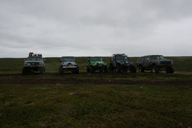 Five modified off-road vehicles parked in a line on a grassy terrain under a cloudy sky.