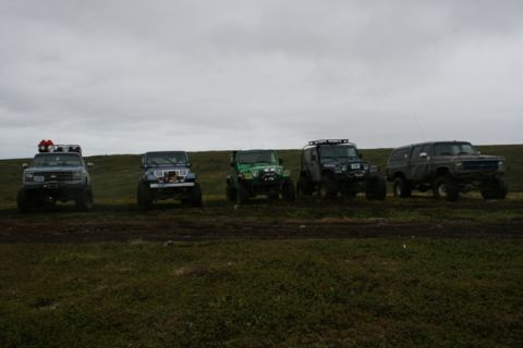 1990 Chevrolet Suburban - Five off-road vehicles, including three Jeeps and two SUVs, are parked in a grassy, muddy field under a cloudy sky. The vehicles are modifi…