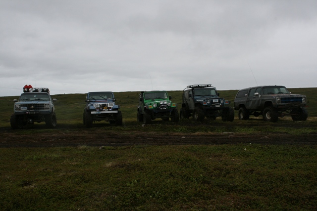 Five modified off-road vehicles parked in a muddy field under a cloudy sky.
