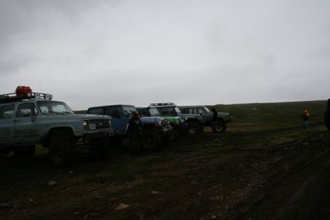1990 Chevrolet Suburban - A line of off-road vehicles, including a light blue Chevrolet Suburban, are parked on a muddy, grassy terrain under an overcast sky. The Su…