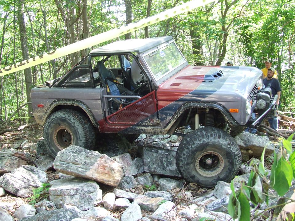 Modified Ford Bronco off-roading over rocks in a forest, with yellow caution tape.