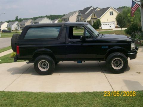 1996 Ford Bronco - Side view of a black 1996 Ford Bronco parked on a concrete driveway in front of a suburban house. The vehicle has a spare tire mounted on t…