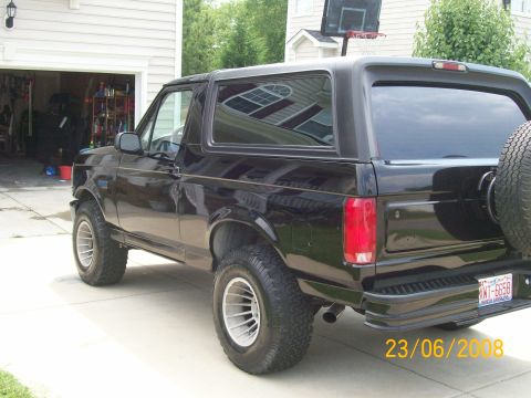 1996 Ford Bronco - The rear passenger side of a black 1996 Ford Bronco is shown parked in a driveway next to a garage. The vehicle has a spare tire mounted on…