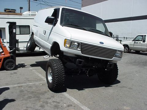2001 Ford E150 - A modified white van with off-road tires is shown from a three-quarter front view, with a pickup truck in the background.