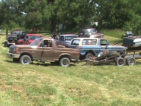 1987 Ford Bronco - A brown 1987 Ford Bronco is parked in a grassy field, towing a trailer with spare tires. Several other trucks and SUVs are visible in the b…