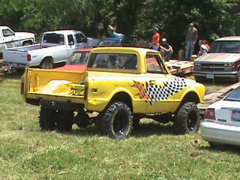 1987 Ford Bronco - A lifted yellow pickup truck with a checkerboard and flame graphic on the side is parked on a grassy field. Other vehicles and people are v…