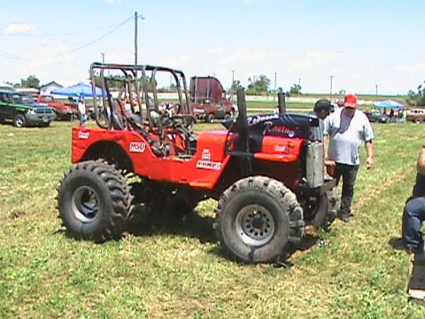 1987 Ford Bronco - A bright red, heavily modified off-road vehicle with oversized tires and a roll cage is parked on a grassy field under a bright blue sky. T…