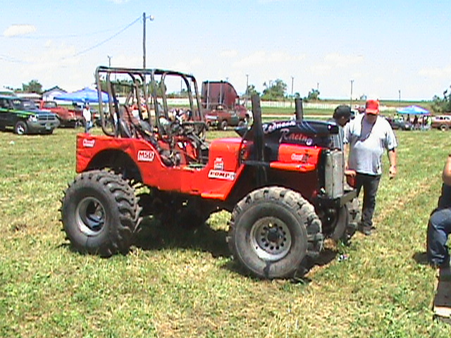 Modified red off-road vehicle with large tires and roll cage parked on grass.