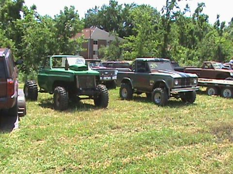 1987 Ford Bronco - Two modified off-road vehicles are parked on a grassy field with trees and buildings in the background. A green SUV with large tires is on…