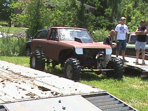 1987 Ford Bronco - A heavily modified, lifted pickup truck with oversized tires is parked on a trailer. Three men stand behind the truck, observing it.