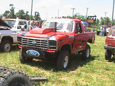 1987 Ford Bronco - A lifted red Ford pickup truck with a large hood scoop and exhaust stack is parked on grass in an outdoor event. A white pickup truck is pa…