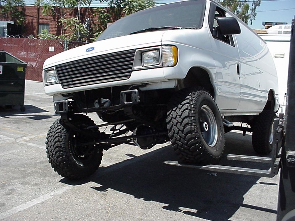 White lifted Ford E-series van on large mud tires, front 3/4 view showing 4x4 suspension