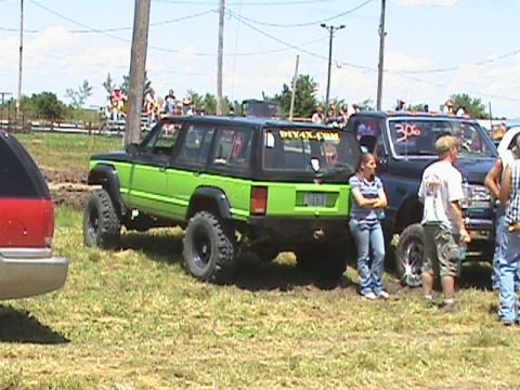 1987 Ford Bronco - A modified green and black Jeep Cherokee XJ with large off-road tires is parked on a grassy field next to a dark blue Ford pickup truck. Se…