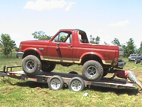 1987 Ford Bronco - A red 1987 Ford Bronco is loaded onto a flatbed trailer, with a person underneath the rear of the vehicle. The Bronco has large off-road ti…