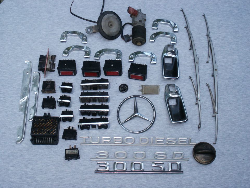 Mercedes-Benz parts including chrome handles, switches, 300 SD badges and star emblem on gray cloth