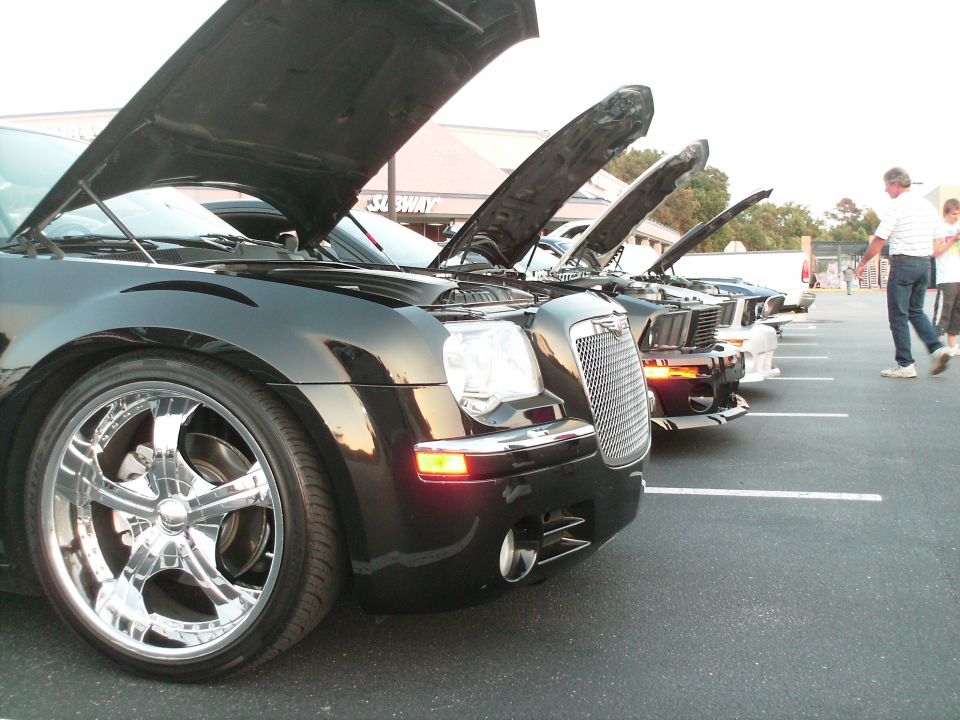 Row of cars with open hoods at a car show, black Chrysler 300 on chrome wheels in front