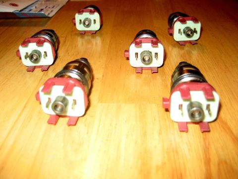 1993 Ford Probe - Seven fuel injectors are laid out on a wooden table. The parts are red and white with silver metal components.