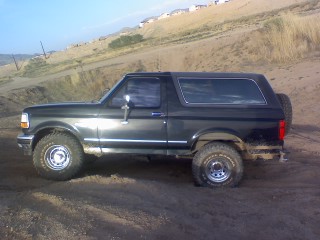 1993 Ford Bronco - A dark green 1993 Ford Bronco is seen from the side, parked on a dirt road with hills in the background.