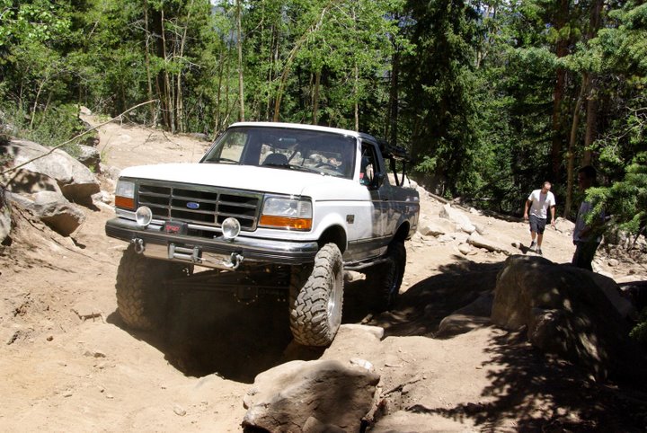 Lifted white Ford Bronco with top off crawling over boulders on a wooded off-road trail