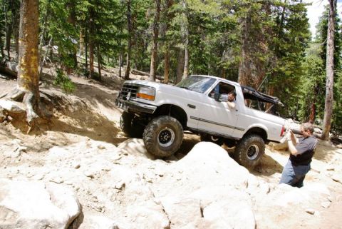 1995 Ford Bronco - A white SUV navigates a challenging rocky terrain on a wooded mountain trail.