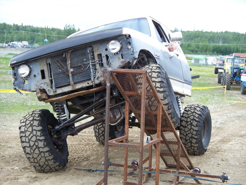 Modified pickup truck with large tires on a metal ramp. Other vehicles in background