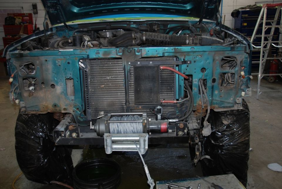 Front of a teal vehicle with hood up, showing engine and radiator. A winch is mounted on the bumper. Tires are covered in black plastic