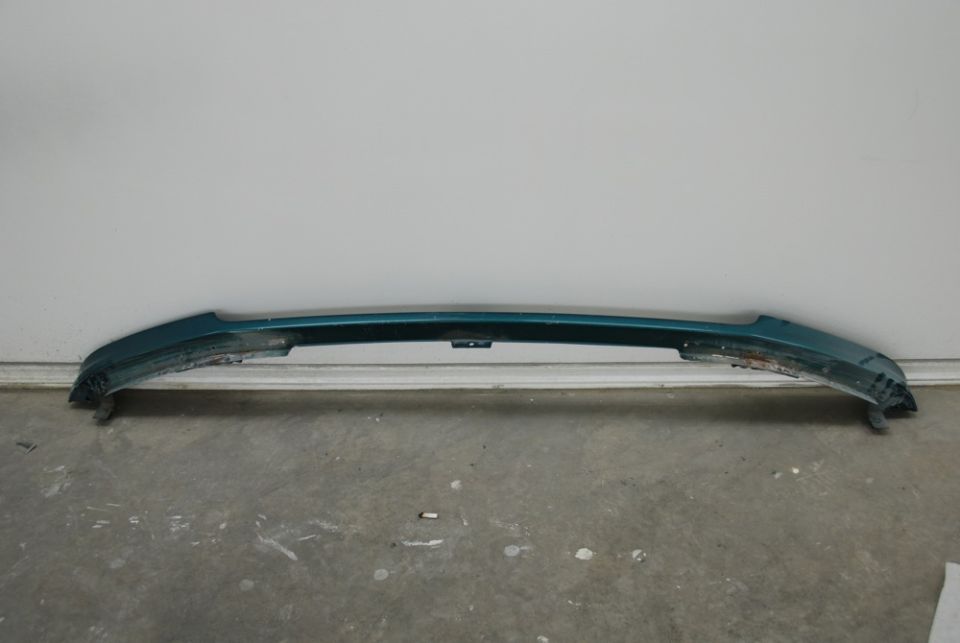 Teal front bumper with damage on the lower edges, mounted on a concrete floor against a white wall