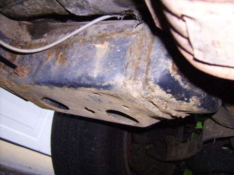 1982 Ford LTD Crown Victoria - Close underbody shot of a rusted frame rail and floor area near a rear wheel, with heavy scale, flaking undercoating, and a torn/rotted sea…