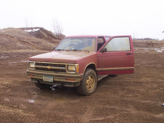 Red pickup truck, possibly a Chevrolet S-10, parked in a muddy field with driver's door open. License plate reads 154-PIE