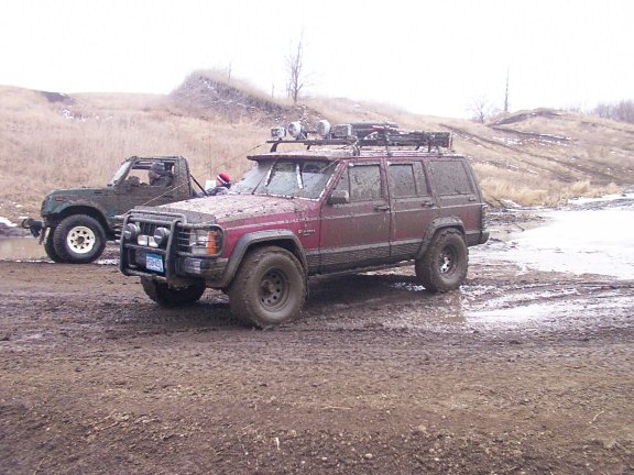 Muddy maroon Jeep Cherokee XJ with roof lights and brush guard on a wet off-road trail