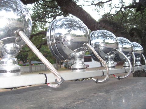 1990 Ford Bronco - A row of chrome KC lights is mounted on a bar, reflecting the surrounding trees and sky.