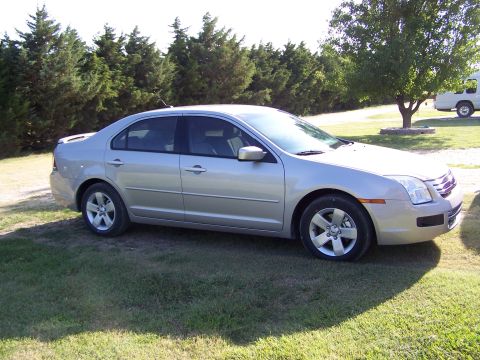 2008 Ford Fusion - A silver sedan is parked on a grassy area with trees and a white RV in the background.