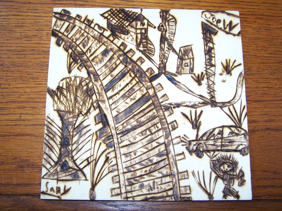 Wood-burned art shows train tracks curving past houses, a car, and figures. Text 'Joe W' and 'Sarl' visible