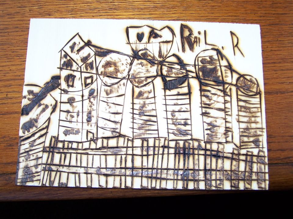 Drawing on paper with wood-burned lines depicting buildings and a fence. Text reads "Rail R."