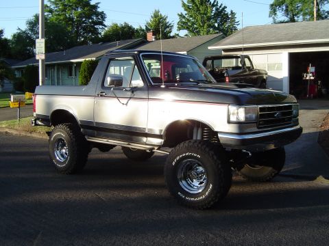 1989 Ford Bronco - A lifted Ford Bronco with off-road tires sits parked in front of a garage, with another vehicle visible inside.