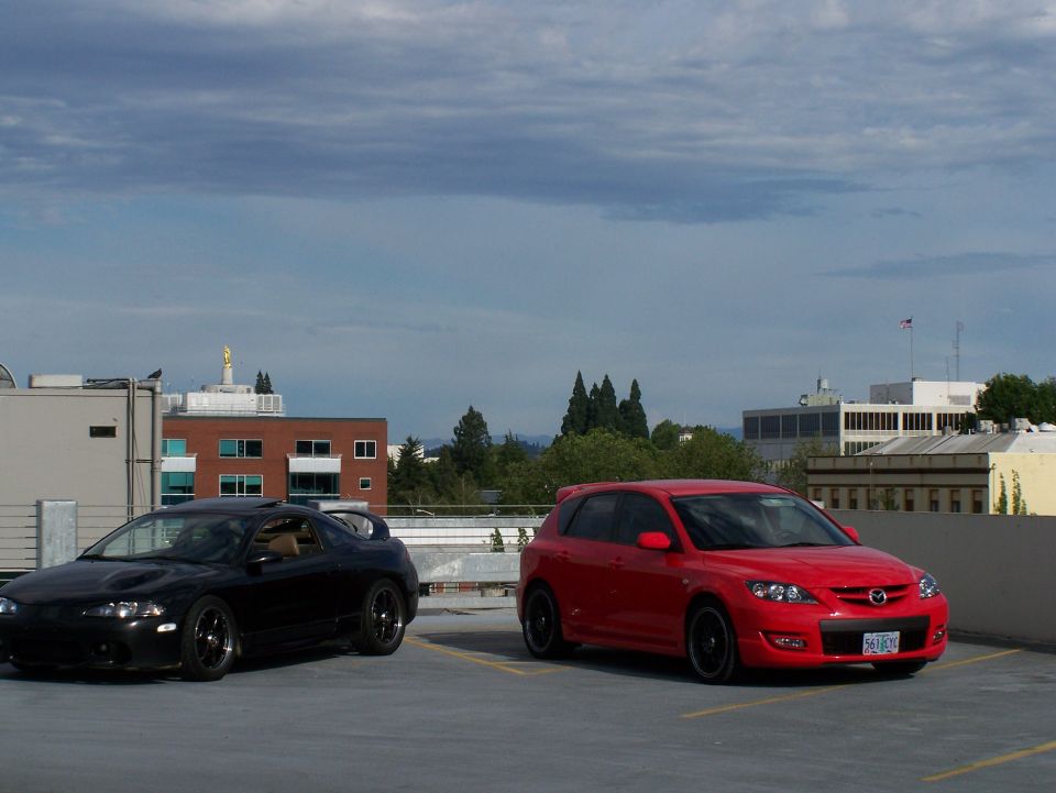 Red 2007 Mazda 3 hatchback parked next to a black coupe on a rooftop