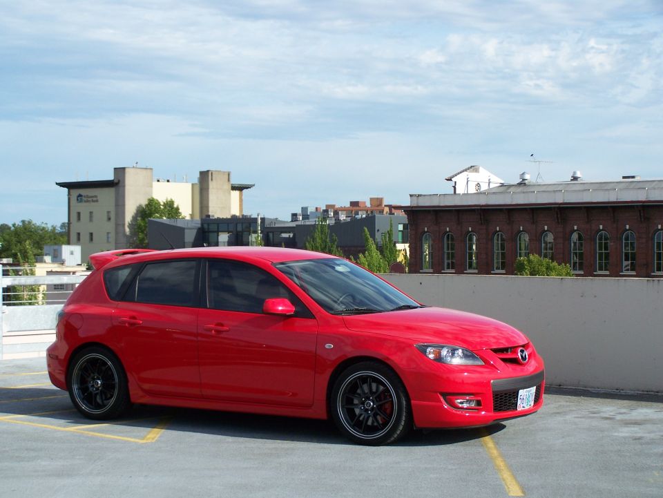 Red 2007 Mazda 3 hatchback with black rims parked on a rooftop, viewed from the front three-quarter angle